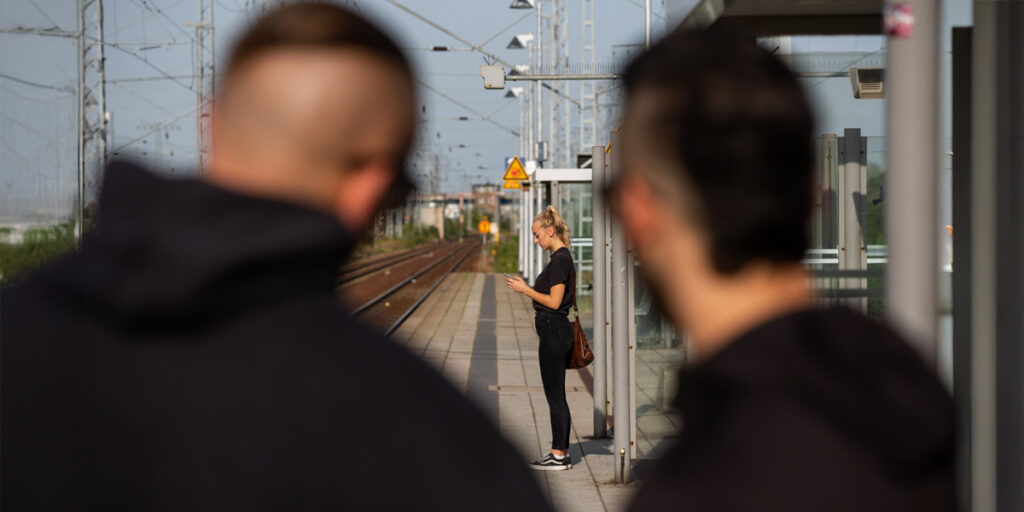 Save Basics_Szenario: Zwei Männer beobachten eine junge Frau, die am Bahnsteig auf ihren Zug wartet
