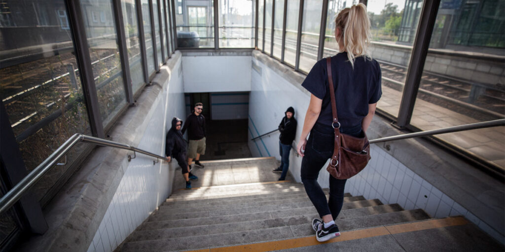 Save Women Ground Survival Szenario: Eine Frau betritt eine Treppe hinab zu einer Bahnunterführung, im unteren Bereich stehen drei bedrohlich wirkende Männer in dunkler Kleidung.